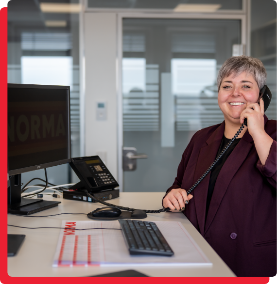 Verwaltung-Statistik-img-2 Eine Person mit kurzen Haaren steht lächelnd in einem Büro und telefoniert. Es gibt einen Schreibtisch mit Computer, Tastatur und Telefon. Im Hintergrund sind Fenster und eine Tür zu sehen.