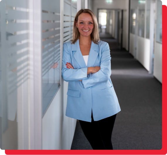 bau-immobilien-expansion-img-1 Auf dem Bild steht eine Frau in einem Büroflur, lehnt sich lächelnd gegen eine Wand. Sie trägt einen hellblauen Blazer und hat die Arme verschränkt.