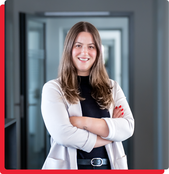 media-kommunikation-marketing-img-2 Eine lächelnde Frau mit langen braunen Haaren steht mit verschränkten Armen in einem Flur. Sie trägt einen hellen Blazer und ein dunkles Oberteil.